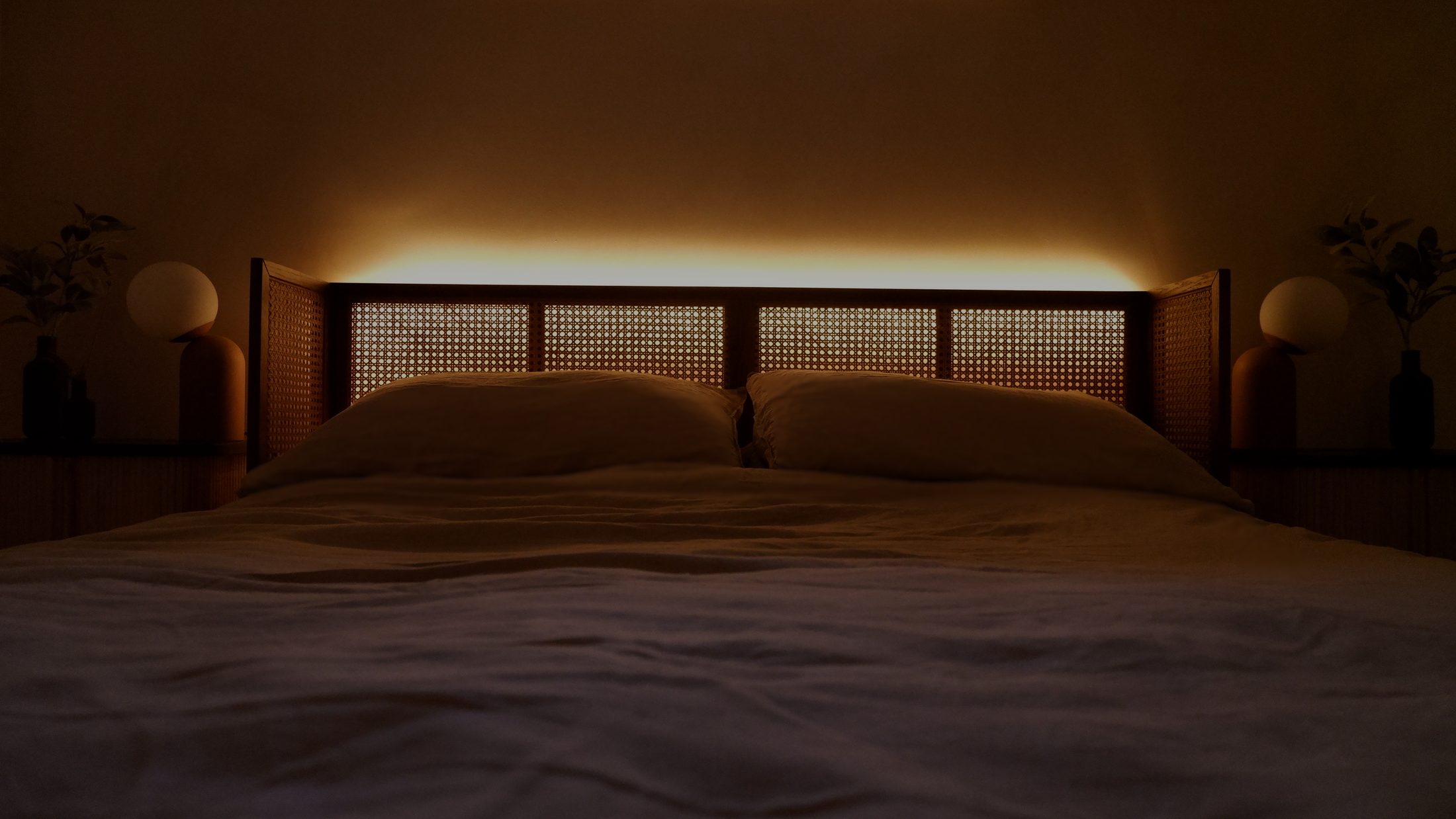 A bed with a cane headboard, backlit with a warm strip of light, globe lamps either side