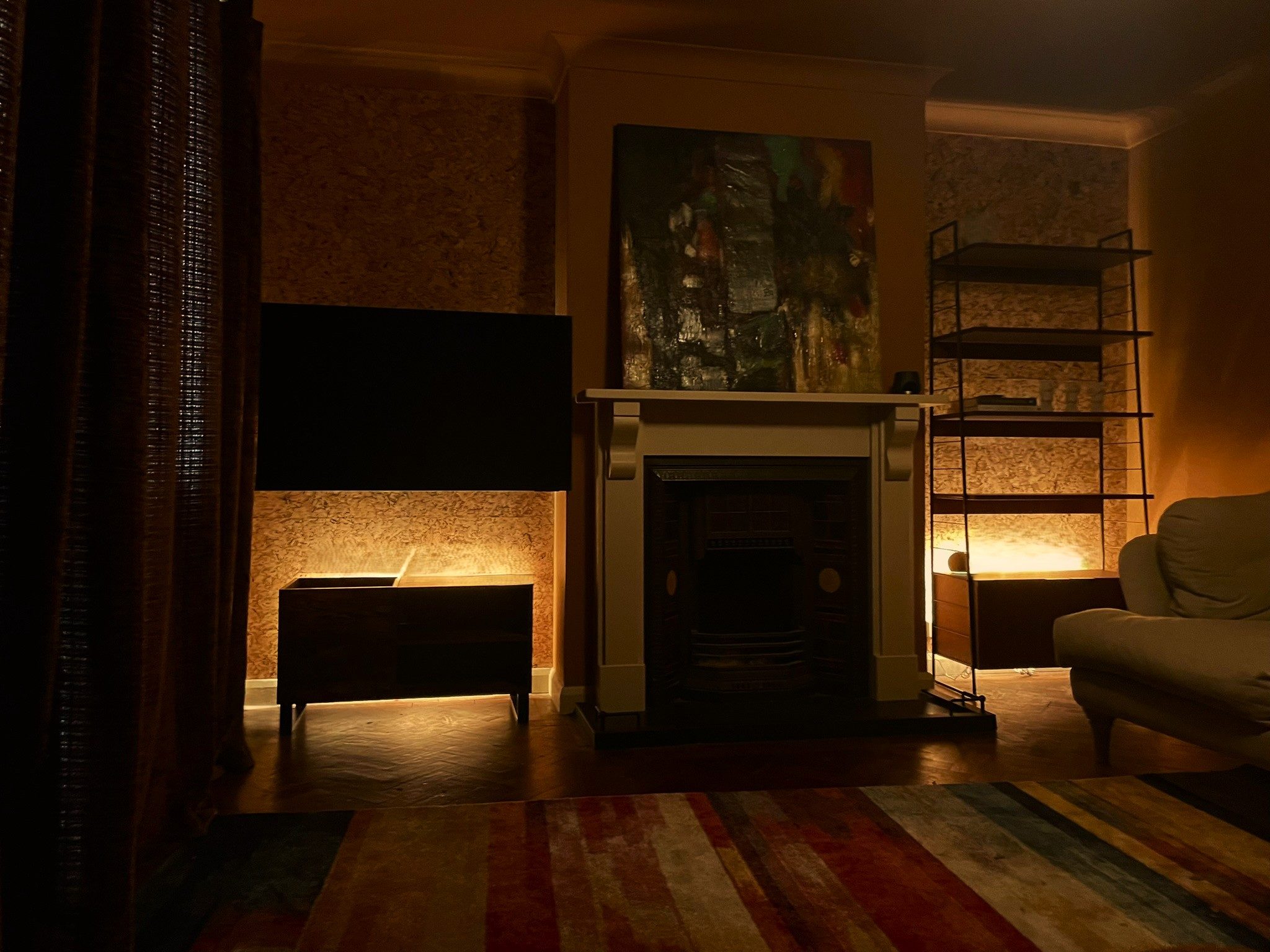 A sitting room at dusk: cork-lined alcoves, a cream fire surround, string shelving and a striped rug